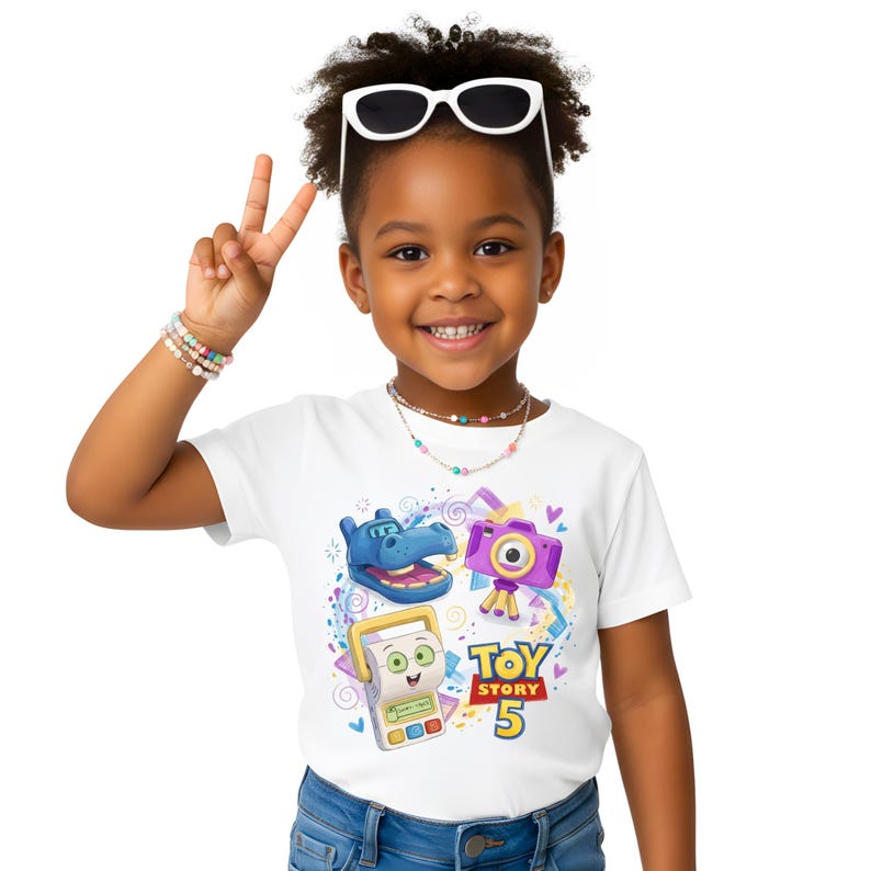 Pu&ograve; includere: Una bambina indossa una maglietta bianca con una grafica colorata di "Toy Story 5". Il design include un ippopotamo blu, una fotocamera viola e un dispositivo giallo. Indossa anche occhiali da sole, una collana e braccialetti.