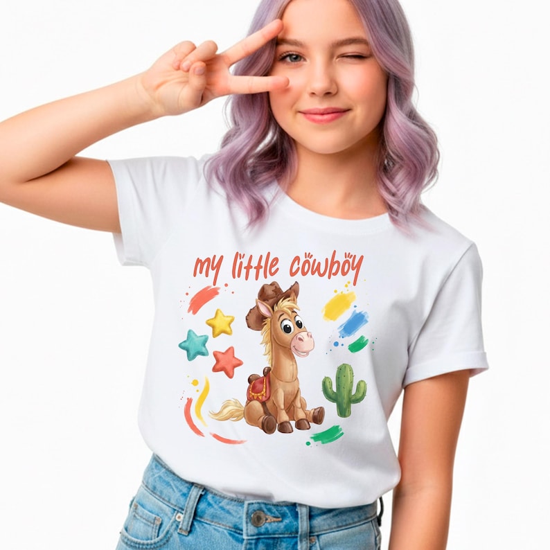 以下が含まれることがあります： 赤い文字で「my little cowboy」と書かれた白いTシャツ。カウボーイハットをかぶった馬の漫画イラストが特徴で、カラフルな星、筆のストローク、サボテンに囲まれています。