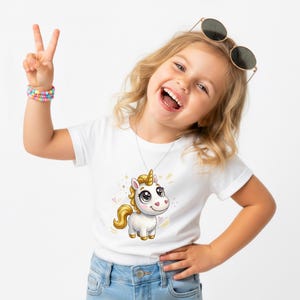 May include: A white t-shirt featuring a cartoon unicorn with a golden horn and mane. The unicorn has large eyes and a smiling face. The shirt is worn by a young person who is smiling and holding up two fingers.