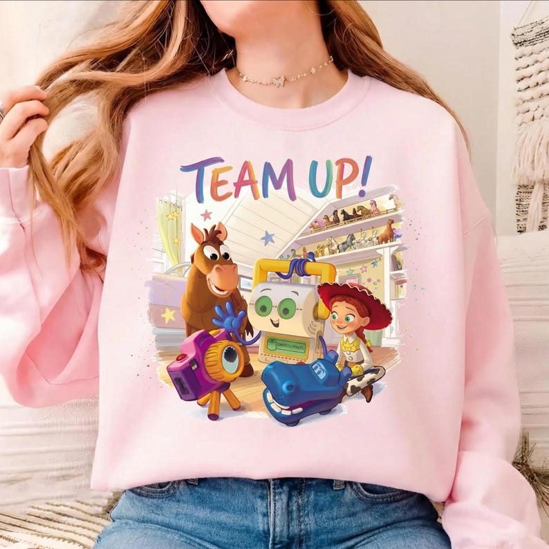 May include: A light pink sweatshirt featuring a colorful graphic of characters from the Toy Story franchise. The phrase "TEAM UP!" is written in rainbow colors across the top. The design includes a horse, a robot, a camera, and other characters.