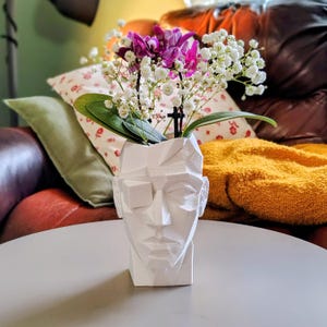 May include: A white, geometric, low-poly style head-shaped vase featuring sharp, angular planes and a faceted surface. The vase contains a floral arrangement of small white baby's breath flowers and vibrant purple orchid blossoms with green leaves.