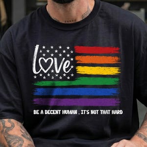 Könnte beinhalten: Ein schwarzes T-Shirt mit einem Grafikdesign einer Flagge mit weißen Sternen und einem Herz, das den Buchstaben O im Wort LOVE ersetzt, gefolgt von horizontalen Regenbogenstreifen und dem Text BE A DECENT HUMAN, IT'S NOT THAT HARD.