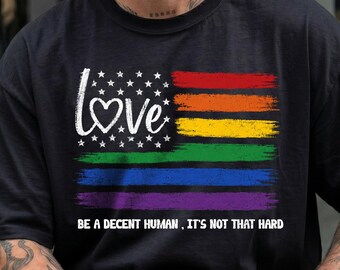 May include: A black t-shirt featuring a graphic design of a flag with white stars and a heart shape replacing the letter O in the word LOVE, followed by horizontal rainbow stripes and the text BE A DECENT HUMAN, IT'S NOT THAT HARD.