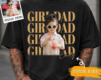 May include: A black short-sleeve t-shirt featuring a repeating gold text pattern that reads GIRL DAD. The center displays a photographic print of a child wearing sunglasses and holding a drink, with the cursive name Emily and a custom name label.