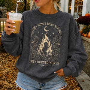 Peut inclure: Un sweat-shirt à col rond gris anthracite foncé avec une impression graphique couleur crème représentant un feu de joie avec la silhouette d'une personne à l'intérieur, entourée de motifs floraux, d'un croissant de lune et du texte "THEY DIDN'T BURN WITCHES THEY BURNED WOMEN."