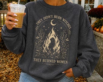 May include: A dark charcoal gray crewneck sweatshirt featuring a cream-colored graphic print of a bonfire with a silhouette of a person inside, surrounded by floral motifs, a crescent moon, and the text "THEY DIDN'T BURN WITCHES THEY BURNED WOMEN."