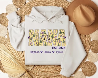 May include: A white hooded sweatshirt featuring a graphic print with the text NANA life EST.2026 and the names Sophia, Emma, and Tyler. The letters in NANA are filled with a yellow pattern of green Christmas trees, purple bows, and snowflakes.