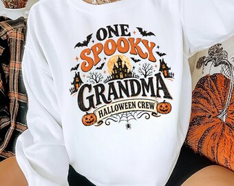 May include: A white long-sleeve sweatshirt featuring a graphic print with the text ONE SPOOKY GRANDMA HALLOWEEN CREW. The design includes orange and black illustrations of a haunted house, a full moon, bats, spider webs, and two jack-o'-lanterns.