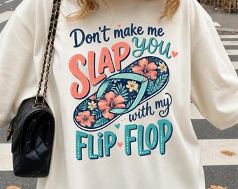 May include: A cream-colored sweatshirt featuring a graphic print of a flip-flop decorated with orange and yellow hibiscus flowers. The text reads, "Don't make me SLAP you with my FLIP FLOP," in a mix of dark blue, coral, and teal stylized lettering.