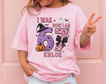 May include: A light pink t-shirt featuring a graphic with the text I WAS NOW I AM KHLOE. The design includes a large purple number 6 with a witch hat and pumpkin, and a pink number 7 with a black bow, surrounded by small ghosts, bats, and spiders.