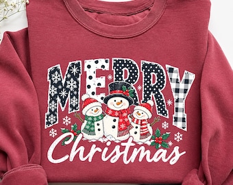 May include: A maroon crewneck sweatshirt featuring the text MERRY in a mix of black and white patterns, including polka dots, snowflakes, and buffalo plaid. Below, the word Christmas is printed in white script above three snowmen.