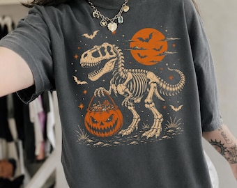 May include: A charcoal gray t-shirt featuring a graphic print of a dinosaur skeleton holding a jack-o'-lantern bucket filled with candy. The design includes an orange full moon, flying bats, and small stars against a dark background.