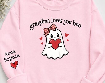May include: A light pink long-sleeve sweatshirt featuring a central graphic of a white ghost with a red and white checkered bow holding a red heart. The text reads "grandma loves you boo". The right sleeve displays the name "Anna Sophia" and a red heart.