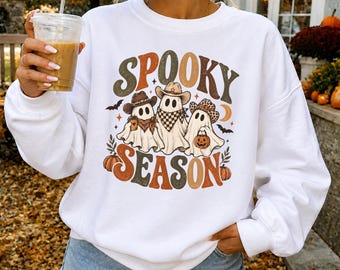 May include: A white long-sleeve sweatshirt featuring a graphic print with the text SPOOKY SEASON. The design includes three ghosts wearing cowboy hats, one holding a coffee cup and another a pumpkin bucket, surrounded by bats and pumpkins.