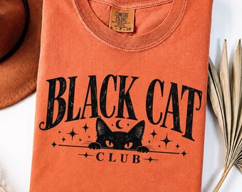 May include: An orange short-sleeved t-shirt with a black graphic print. The text reads BLACK CAT CLUB. The graphic features a black cat face with glowing eyes peeking over a line, surrounded by small stars and a crescent moon. The shirt has a crew neck.