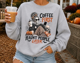 May include: A light gray crewneck sweatshirt featuring a graphic print of a skeleton with a messy bun, sunglasses, and leopard print pants. The skeleton holds a coffee cup and a smartphone. Text reads: COFFEE FIRST HAUNT PEOPLE LATER.