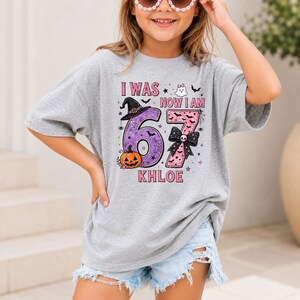 May include: A heather gray t-shirt featuring a Halloween-themed graphic with the text I WAS 67 NOW I AM KHLOE. The design includes a purple number 6 with a witch hat and pumpkin, a pink number 7 with a black bow, a ghost, bats, and spiderwebs.