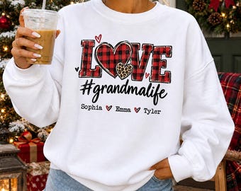 May include: A white long-sleeve sweatshirt featuring the word LOVE in red and black buffalo plaid, with a leopard print heart inside the O. Below is the text #grandmalife and the names Sophia, Emma, and Tyler separated by small red hearts.