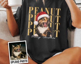 May include: A black short-sleeve t-shirt featuring a graphic of a calico cat wearing a red and white Santa hat. The text "PEANUT" is repeated in gold letters behind the cat, with "Merry Christmas" written in white script below the cat.