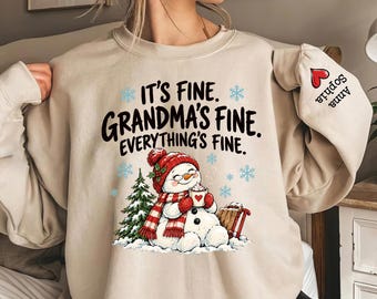 May include: A beige crewneck sweatshirt featuring the black text IT'S FINE. GRANDMA'S FINE. EVERYTHING'S FINE. A snowman wearing a red hat and scarf holds a mug next to a tree and sled. A red heart and the names Sophia and Anna are on the sleeve.