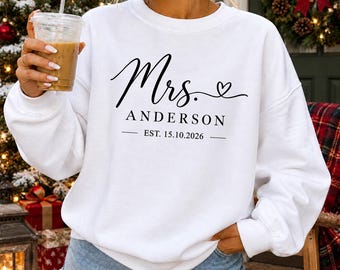 May include: A white long-sleeve crewneck sweatshirt featuring black text that reads Mrs. ANDERSON and EST. 15.10.2026 with a small heart graphic. The garment is made of a soft fabric and has a relaxed fit with ribbed cuffs and a ribbed neckline.