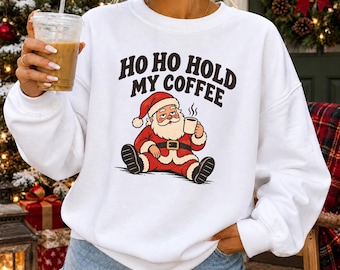 May include: A white crewneck sweatshirt featuring a graphic of Santa Claus sitting and holding a coffee mug. Above the illustration, the text reads HO HO HOLD MY COFFEE in bold black letters. The sweatshirt has long sleeves and a ribbed neckline.