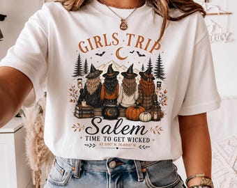 May include: A white t-shirt featuring a graphic of four figures in witch hats sitting before a forest and mountains. The text reads GIRLS TRIP, Salem, TIME TO GET WICKED, and 42.5197 N, 70.8955 W, accompanied by pumpkins, a cauldron, and books.