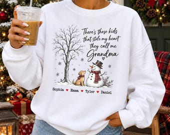 May include: A white long-sleeve sweatshirt featuring a graphic of a bare tree, a golden dog, and a snowman wearing a plaid scarf. The text reads: There's these kids that stole my heart they call me Grandma, followed by the names Sophia, Emma, Tyler, and Daniel.