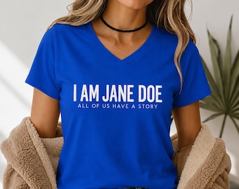May include: A royal blue V-neck t-shirt featuring the white text I AM JANE DOE and the smaller text ALL OF US HAVE A STORY printed on the front. The shirt is worn with a tan fuzzy jacket and blue denim jeans with a visible metal button.