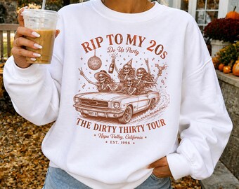 May include: A white crewneck sweatshirt featuring a brown graphic print of skeletons in a convertible car. The text reads RIP TO MY 20s, Do Us Party, THE DIRTY THIRTY TOUR, Napa Valley, California, and EST. 1996. The design includes a disco ball and confetti.