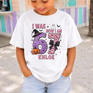 May include: A white short-sleeved t-shirt featuring a graphic with the text I WAS NOW I AM KHLOE and the number 67. The design includes a purple number six with a witch hat and pumpkin, and a pink number seven with a black bow and skull.