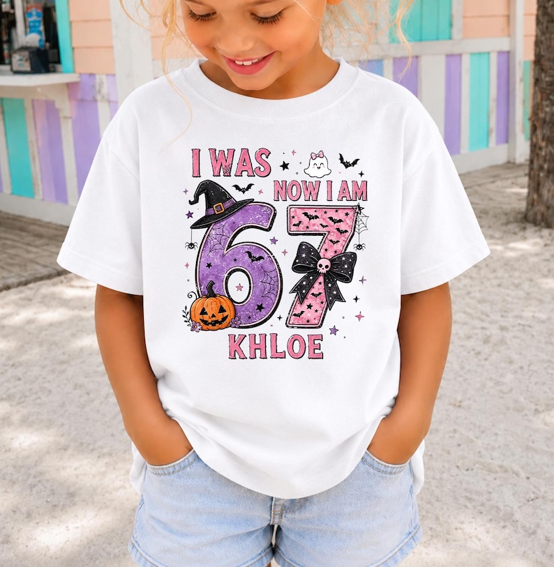May include: A white short-sleeved t-shirt featuring a graphic with the text I WAS NOW I AM KHLOE and the number 67. The design includes a purple number six with a witch hat and pumpkin, and a pink number seven with a black bow and skull.