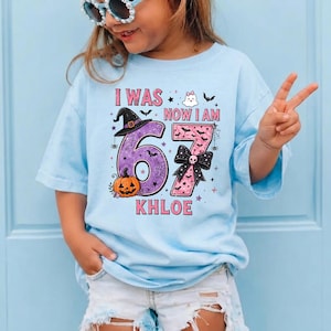 May include: A light blue t-shirt featuring a graphic with the text I WAS NOW I AM and the name KHLOE. The design includes a purple number 6 with a witch hat and pumpkin, and a pink number 7 with a black bow, ghost, and bat illustrations.