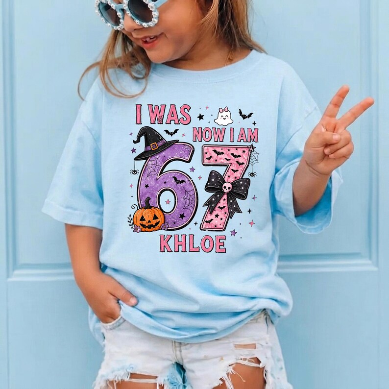 May include: A light blue t-shirt featuring a graphic with the text I WAS NOW I AM and the name KHLOE. The design includes a purple number 6 with a witch hat and pumpkin, and a pink number 7 with a black bow, ghost, and bat illustrations.