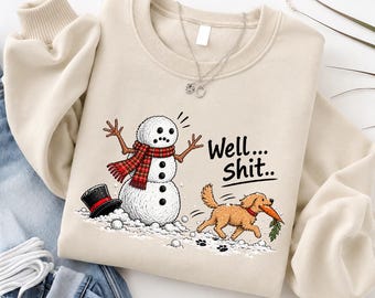 May include: A beige crewneck sweatshirt featuring a graphic of a snowman with a red plaid scarf and a black top hat, and a golden dog running away with the snowman's carrot nose in its mouth. The text "Well... Shit.." is printed in black.