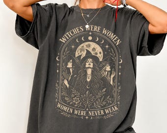 May include: A charcoal gray short-sleeve t-shirt featuring a beige graphic print of three figures in witch hats under a full moon. The text reads "WITCHES WERE WOMEN" at the top and "WOMEN WERE NEVER WEAK" at the bottom, framed by botanical elements.
