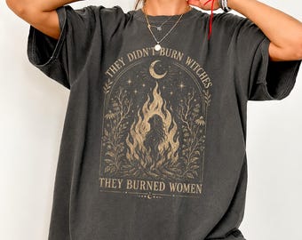 May include: A charcoal gray short-sleeve t-shirt featuring a beige graphic print of a person standing in flames under a crescent moon. The text reads, "THEY DIDN'T BURN WITCHES THEY BURNED WOMEN," framed by botanical illustrations and stars.