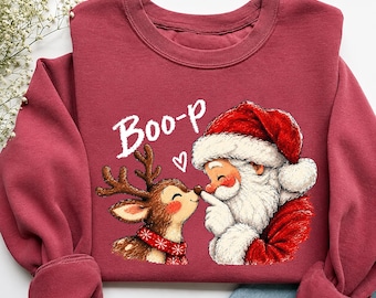 May include: A maroon long-sleeve sweatshirt featuring a graphic print of Santa Claus touching the nose of a reindeer. The text "Boo-p" is printed in white above the characters, accompanied by a small white heart icon between them.