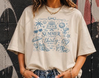 May include: A beige short-sleeve t-shirt featuring a blue graphic print with the text The SUMMER I TURNED thirty EST. 1996. The design includes illustrations of a birthday cake, palm tree, disco ball, beach umbrella, flip-flops, and ice cream.