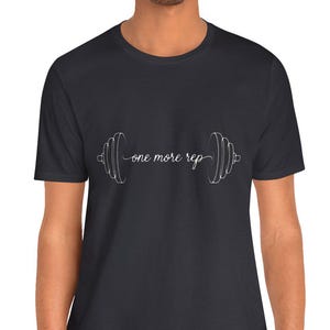 Puede incluir: Camiseta gris oscuro con el texto "one more rep" en blanco, en cursiva, y un gráfico de barra con pesas. La camiseta es de cuello redondo y la lleva una persona.