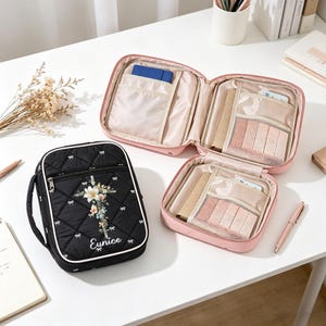 May include: Black quilted travel case with a floral cross design and the name "Eunice". The case has a white trim and a handle. A pink, open, square travel case with multiple compartments is also visible.