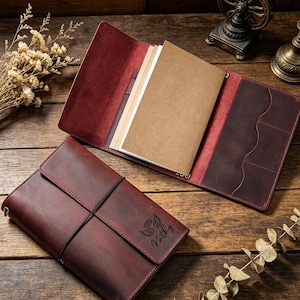 May include: Two burgundy leather journals on a wooden surface, one open and one closed. The open journal shows paper inserts. The closed journal has a black elastic closure and the name "Kathy" with angel wings engraved on the cover.