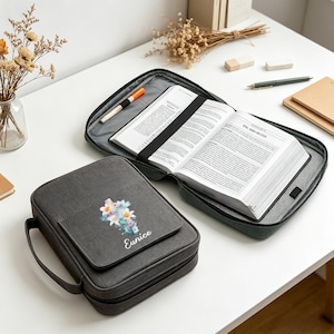 May include: Two grey book covers with floral designs and the name "Eunice" are displayed. One is open, revealing pages held by a black elastic band. A pen and a book are visible inside. The other cover is closed, with a handle.