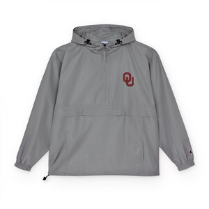 May include: A gray Champion anorak jacket with a hood and a quarter-zip closure. The jacket features a red and white "OU" logo on the chest and a small Champion logo on the sleeve. The jacket has a front pocket and adjustable drawstrings.