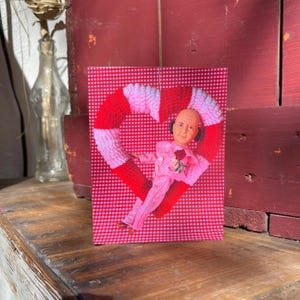 Op de afbeelding: Een wenskaart met een vintage pop in een roze pak, met een roos, in een rood-wit gebreid hart. De achtergrond is een rood-wit ruitpatroon. De kaart wordt gepresenteerd op een houten oppervlak.