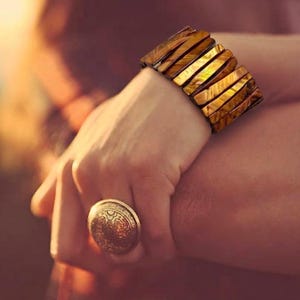 May include: A gold-toned bracelet with rectangular segments and a matching ring with an embossed design. The jewelry is worn on a wrist and hand, with a blurred background.