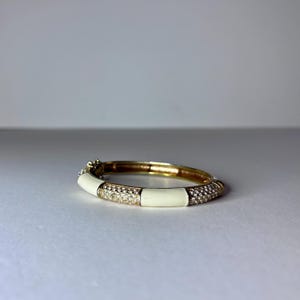 May include: A gold-toned bangle bracelet with white enamel sections and sections encrusted with small, clear crystals. The bracelet is closed and lies on a white surface.