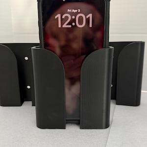 Könnte beinhalten: Schwarzer Telefonhalter mit geschwungenem Design, der ein Smartphone mit der Anzeige "Fri Apr 3 12:01" hält. Der Halter besteht aus einem mattschwarzen Material. Mehrere Halter sind sichtbar, was auf ein Mehrfachprodukt hindeutet.