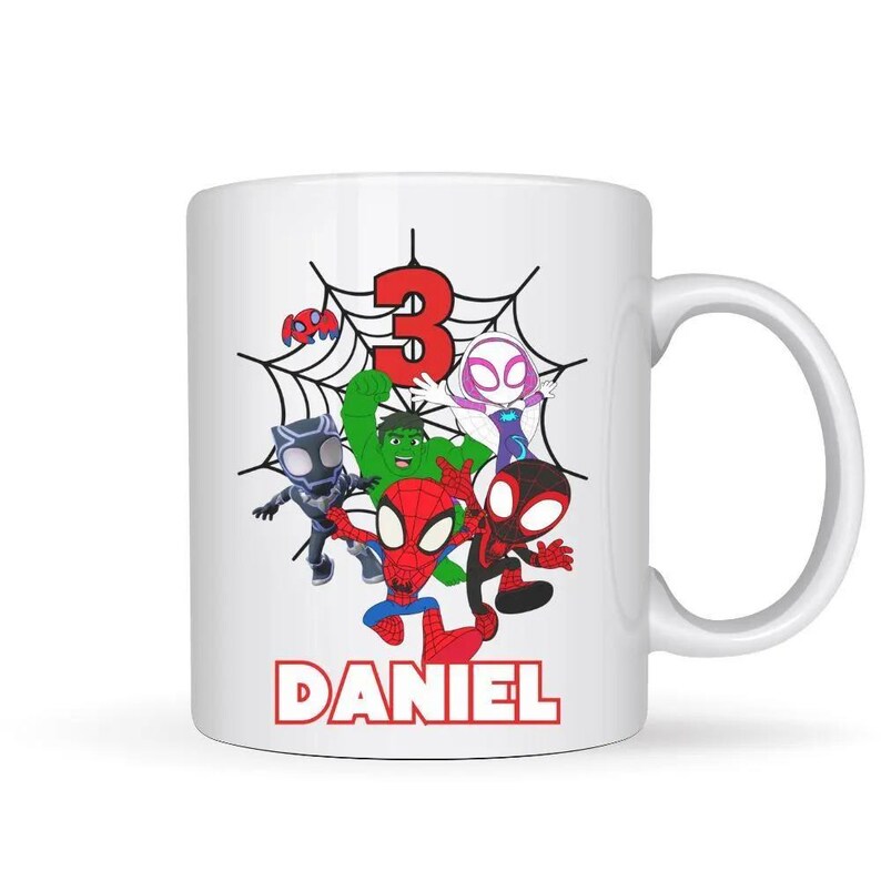 May include: White ceramic mug featuring a colorful graphic of cartoon superheroes, a red number 3, and the name "DANIEL" in red. The superheroes include Spider-Man, Hulk, and others, set against a spiderweb background. A fun, personalized gift.
