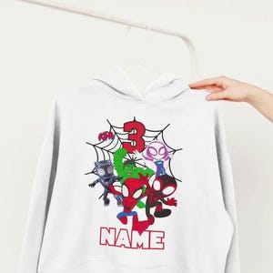 May include: White hooded sweatshirt featuring a colorful graphic of several cartoon superheroes, a red number 3, and the word "NAME" in red block letters. The design is set against a spiderweb background.